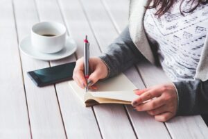 a close up of a woman writing in a journal beside her cup of coffee